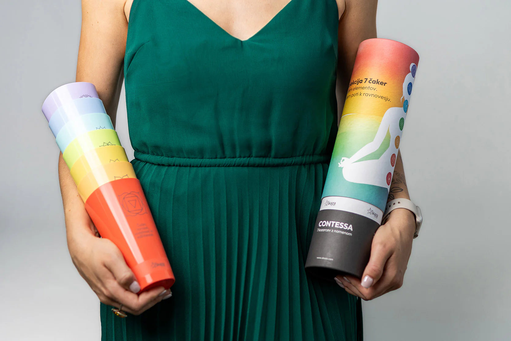 Frau im grünen Kleid hält den kompletten Satz der sieben farbigen CONTESSA Chakra-Becher sowie die passende Verpackungsröhre mit Chakra-Motiv in den Händen.