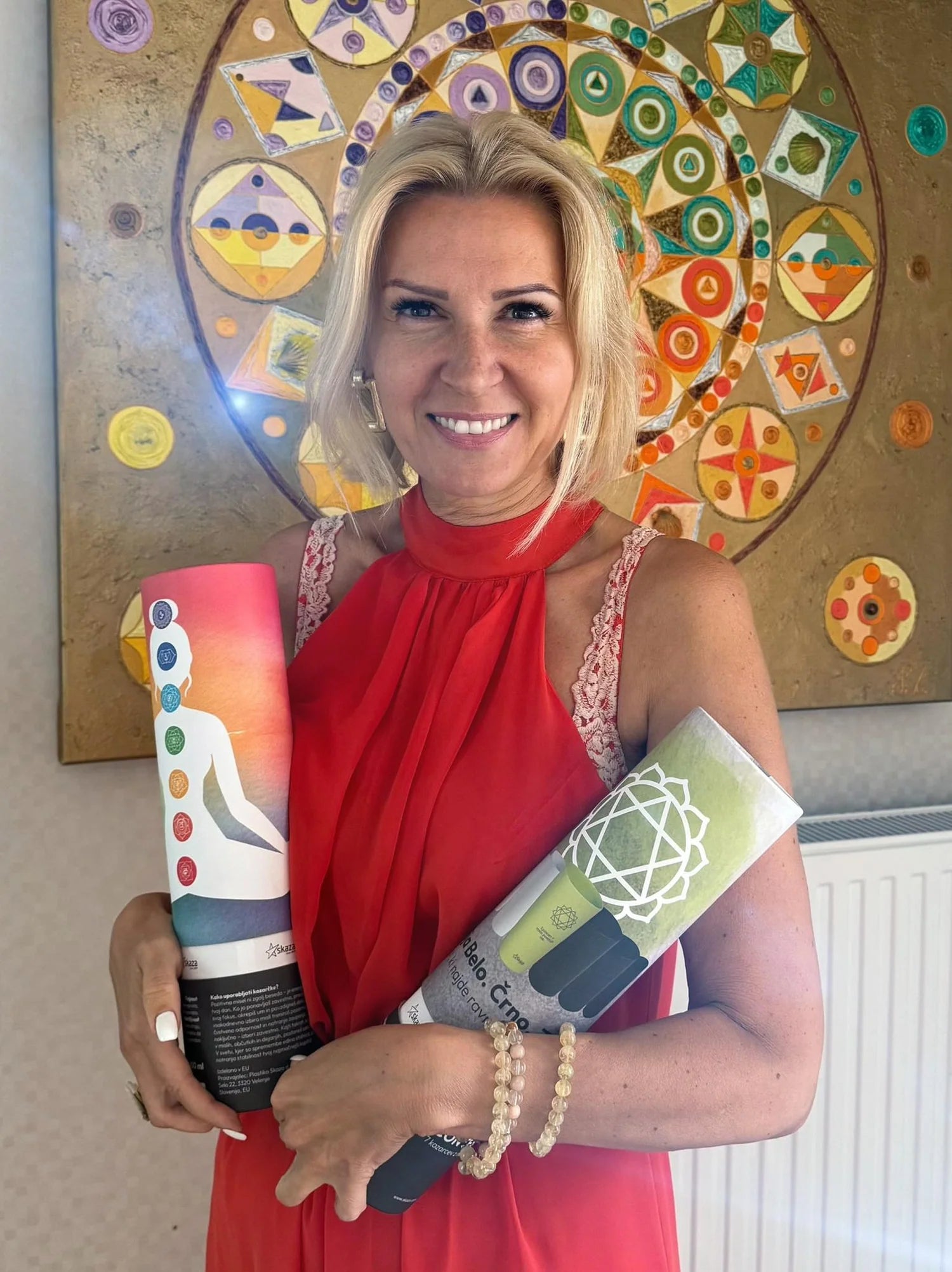 Eine Frau in rotem Kleid hält zwei CONTESSA Verpackungsröhren mit Chakra-Bechern vor einem farbenfrohen Mandala-Hintergrund.