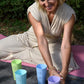 Frau in beigem Kleid sitzt auf einer Yogamatte im Freien und ordnet farbige CONTESSA Chakra-Becher, die jeweils mit Chakra-Symbolen und Affirmationen bedruckt sind.