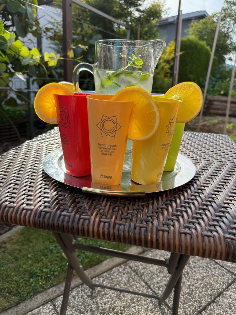 Bunte Chakra-Becher mit Orangenscheiben und einer Karaffe mit Erfrischungsgetränk stehen auf einem Tablett auf einem Gartentisch.