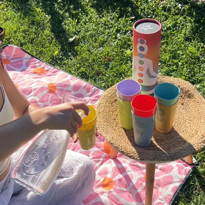 Person gießt beim Picknick Wasser aus einer Glaskaraffe in einen gelben Chakra-Becher, auf dem Tischchen stehen weitere bunte Becher und eine Geschenkverpackung.