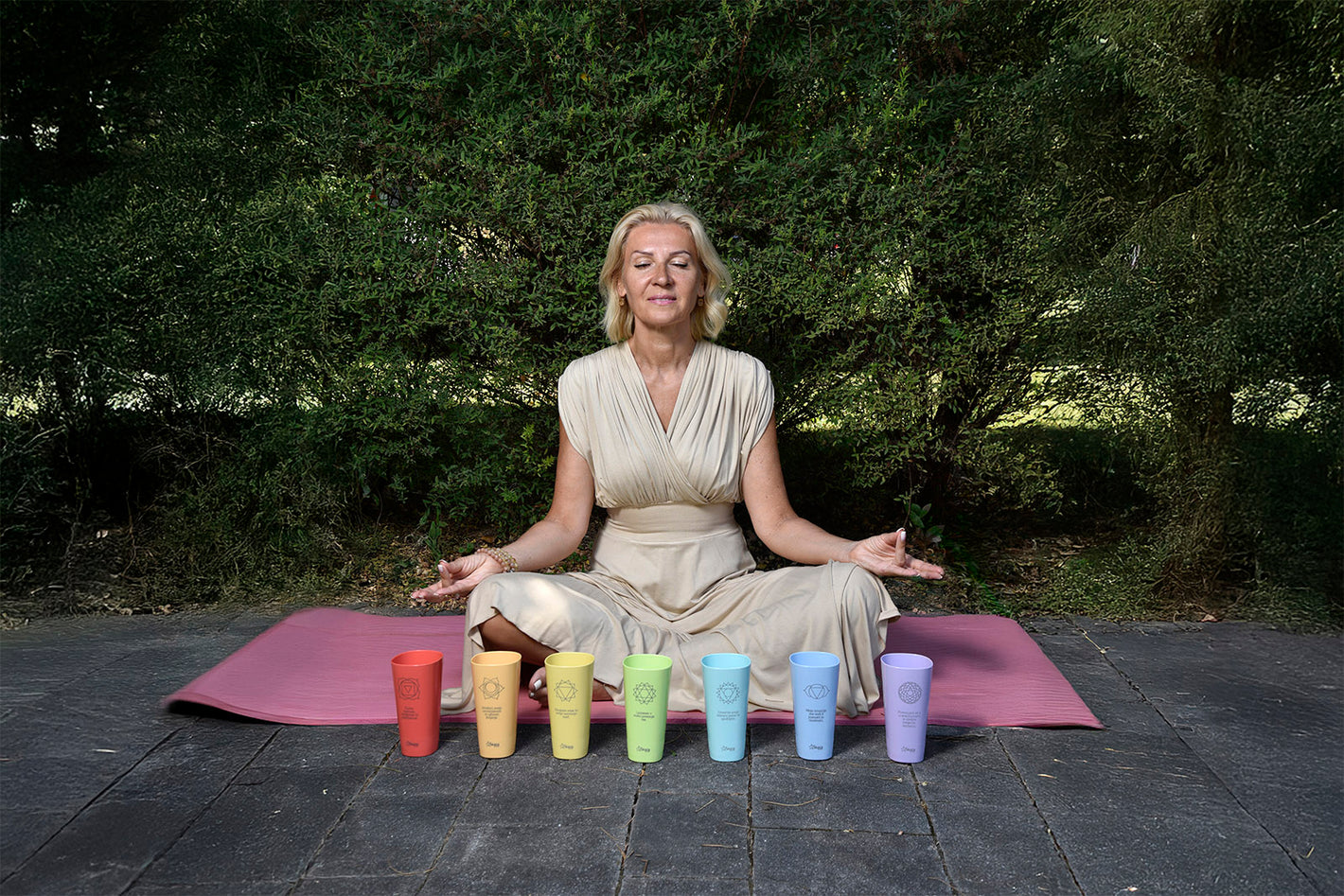 Frau sitzt meditierend auf einer Yogamatte, vor ihr stehen sieben SKAZA bunte Becher mit Chakra-Symbolen und Affirmationen.